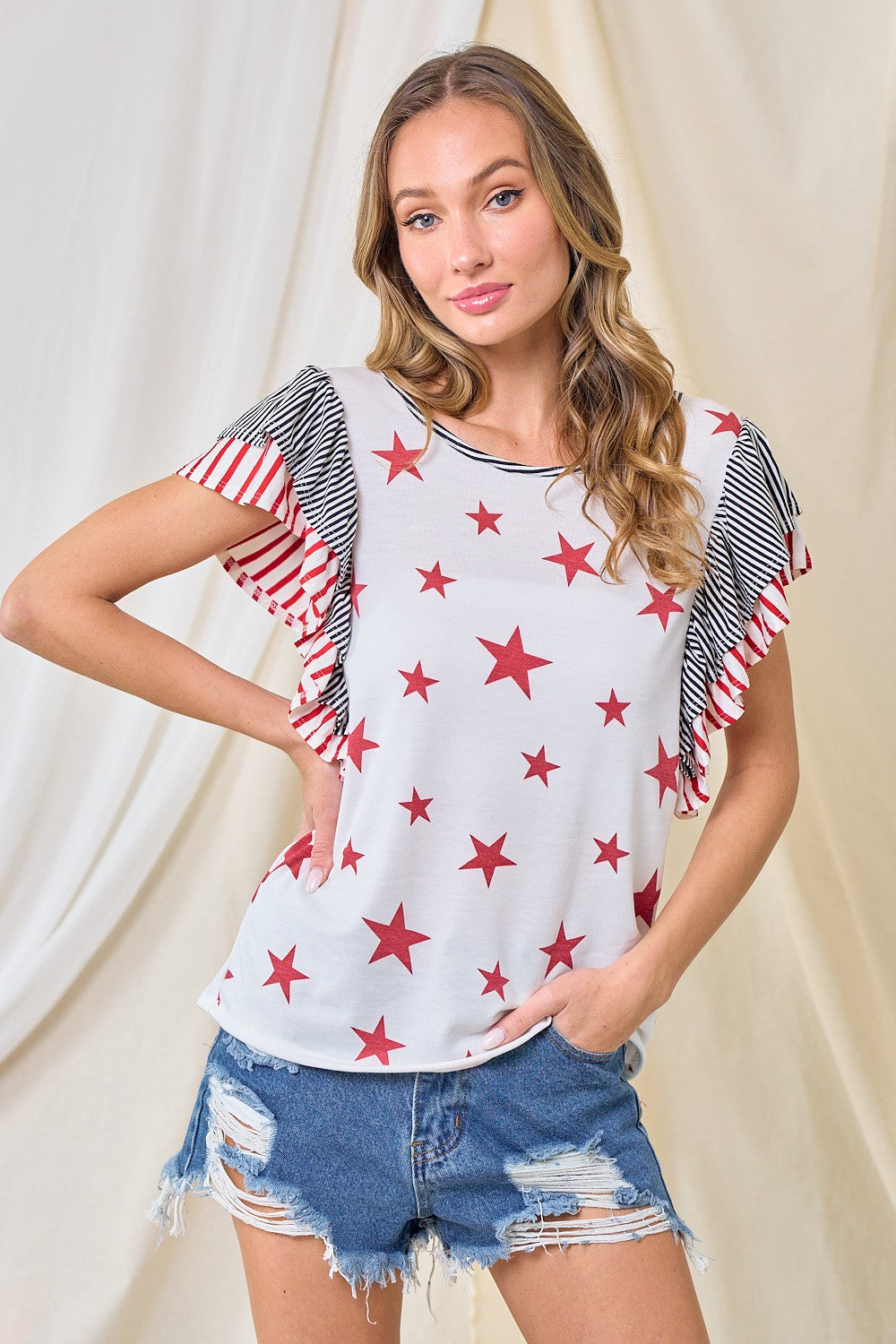 Woman wearing a white t-shirt with red star patterns and denim shorts against a beige curtain background