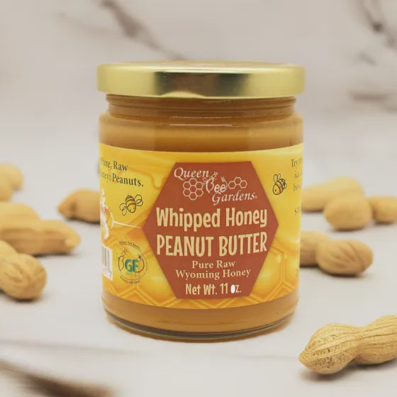 Jar of Queen Bee Gardens Whipped Honey Peanut Butter with peanuts around on a light background