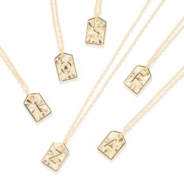 Five gold initial necklaces with textured, engraved letter pendants on delicate chains, arranged diagonally on a clean white background.