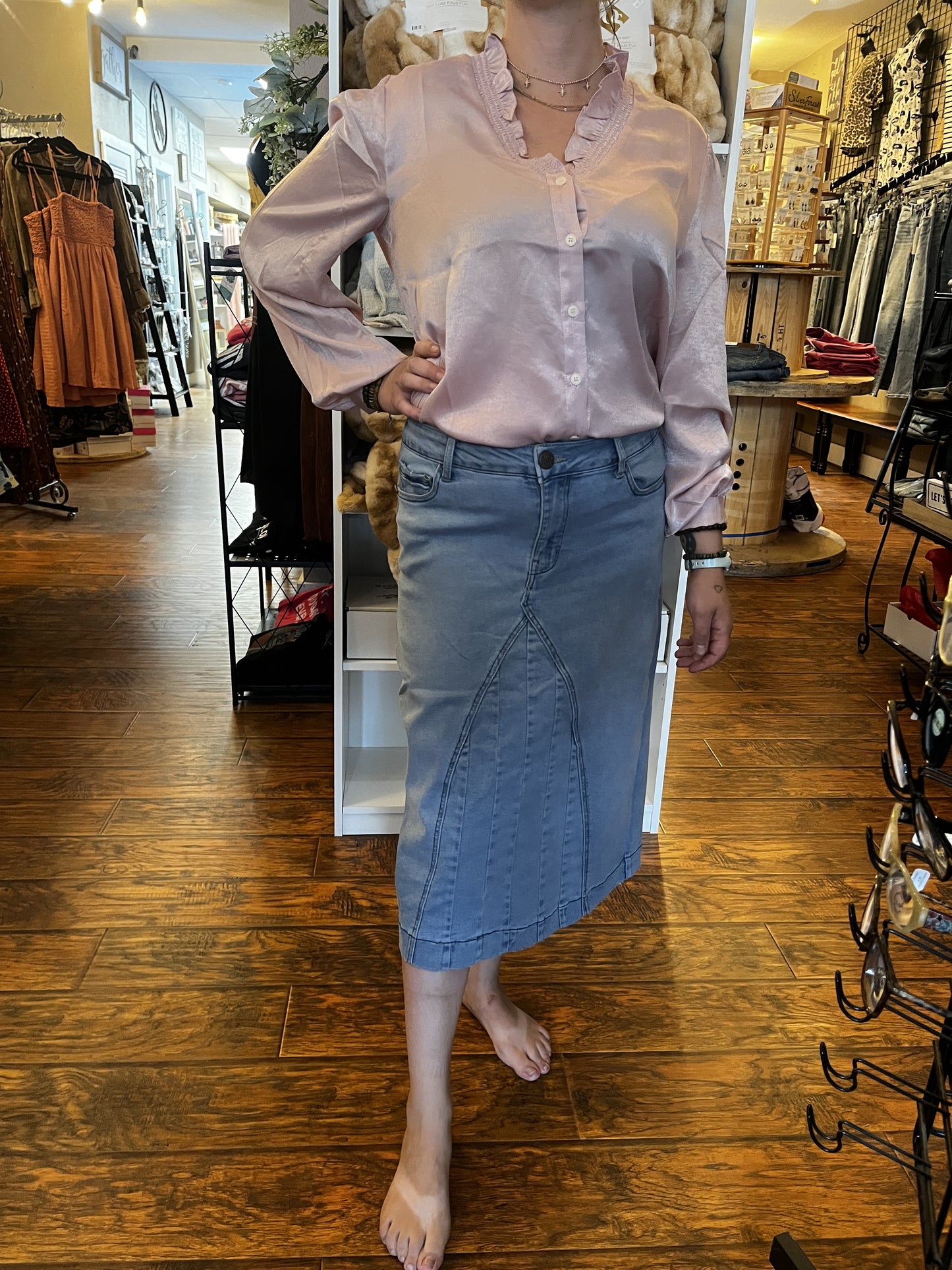 A woman modeling a light pink satin long-sleeve blouse with ruffled trim and a light blue denim midi skirt, standing in a clothing store.