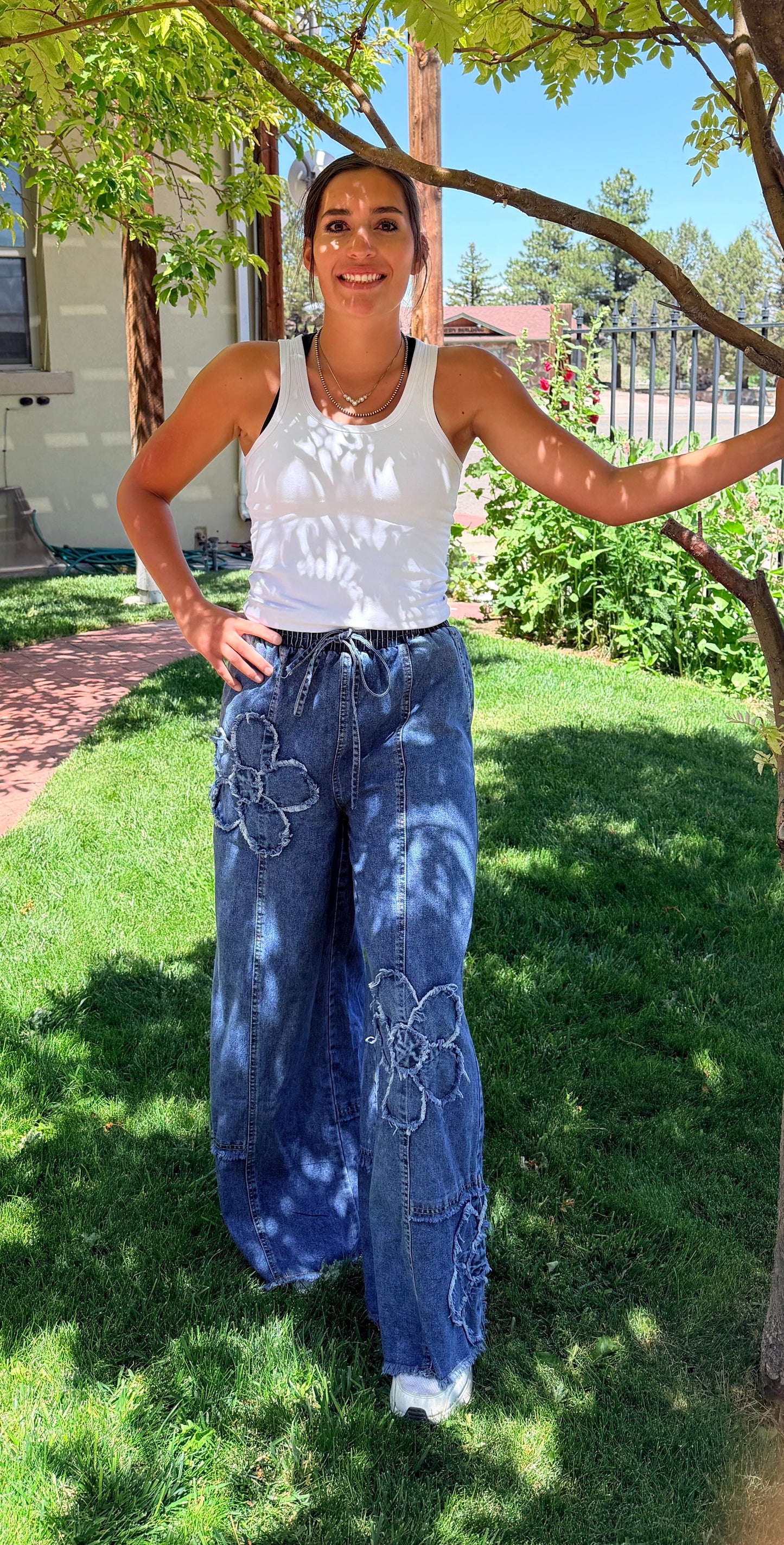Woman wearing a white sleeveless top and blue floral pants standing in a grassy area with trees.